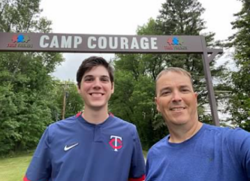 A young man and his uncle wear blue t-shirts and smile in front of a sign outside that says Camp Courage
