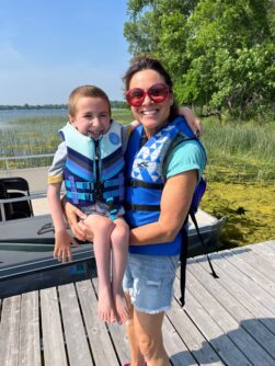 A woman in a life-vest holds a small child also in a life vest in front on a deck in front of a boat on a lake